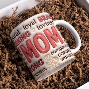 May include: White ceramic mug with the word "MOM" in large, pink letters. The mug is decorated with words like "beautiful," "loving," and "strong." The mug is nestled in brown packing material inside a white box.
