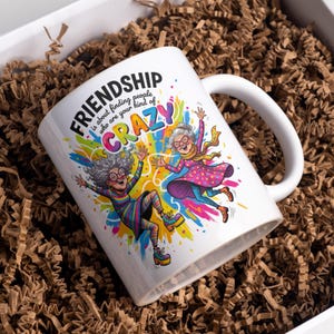 May include: White ceramic mug with a colorful illustration of two elderly women jumping for joy. The mug features the text "FRIENDSHIP is about finding people who are your kind of CRAZY."