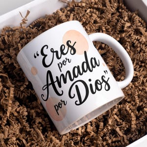 May include: White ceramic mug with the text "Eres por Amada por Dios" in black script, accented with peach-coloured circles. The mug is nestled in brown paper filler inside a white box, ideal for gifting.