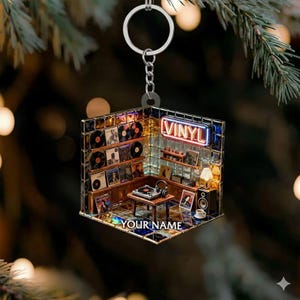 May include: A vinyl record shop keychain with a miniature interior scene. The keychain features a turntable, records, and the word "VINYL" in neon. The bottom of the keychain has the text "YOUR NAME".