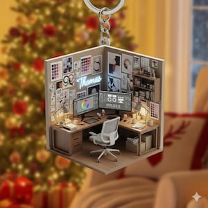 May include: A miniature, three-dimensional ornament depicting a graphic designer's workspace. The ornament features a desk with two computer monitors, a chair, and various design elements. The name "Thomas" is displayed in neon. The background includes a Christmas tree.