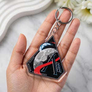 May include: Clear acrylic keychain with a space-themed design. The keychain features a black triangle with a moon, Earth, and a red stripe. The names GLOVER, WISEISM, and KOCH HANSEN are printed on the keychain. A silver keyring is attached.