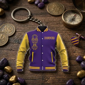 May include: A purple and yellow jacket-shaped keychain with the word "DEUCE" and Greek letters. The keychain is on a wooden surface with coins, a compass, and decorative stones. The jacket has yellow sleeves and trim.