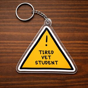 May include: A triangular acrylic keychain featuring a yellow warning sign design with a black border and an exclamation point. The text TIRED VET STUDENT is printed in black capital letters in the center. It includes a silver metal ring and chain.