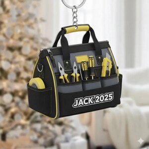 May include: A gray and black toolbox-shaped ornament with yellow accents, featuring miniature tools in the pockets. The text "JACK 2025" is printed on the front. The ornament hangs from a silver chain.