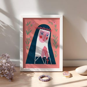 Puede incluir: Una impresión artística enmarcada que presenta una ilustración estilizada de una monja con las manos juntas. La obra de arte tiene un fondo rosa con detalles florales y un marco blanco. La monja lleva un velo negro y una corona de espinas.
