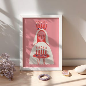 May include: A framed art print featuring a stylized illustration of a figure with a crown and holding candles. The artwork has a pink background and is framed in white. A purple bracelet, a gold ring, and a stone are in the foreground.