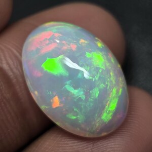 Puede incluir: Una piedra preciosa de ópalo pulida, de forma ovalada. El ópalo muestra un color base blanco lechoso con destellos vibrantes de verde, naranja y rojo. La gema se sostiene en una mano, mostrando su juego de colores iridiscente.