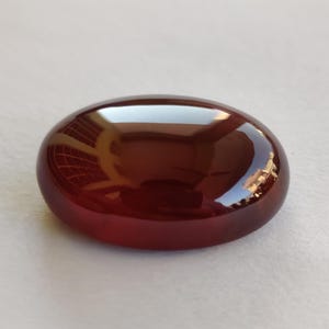 May include: A polished, oval-shaped gemstone with a deep reddish-brown hue. The surface reflects light, creating highlights and shadows. The gemstone is smooth and appears to be a cabochon, suitable for jewelry.