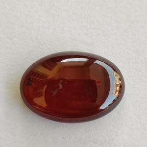 Könnte beinhalten: Ein polierter, oval geformter Edelstein in einem tiefen Rotbraun. Die Oberfläche ist glatt und reflektierend, wobei sich das Licht auf der polierten Oberfläche spiegelt. Der Edelstein liegt vor einem weißen Hintergrund.