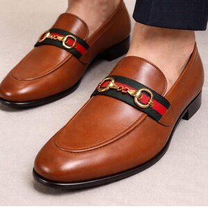 Puede incluir: Mocasines de cuero marr&oacute;n con hebilla dorada y detalle de rayas verdes y rojas. Los zapatos tienen un dise&ntilde;o cl&aacute;sico con punta redondeada y tac&oacute;n bajo.