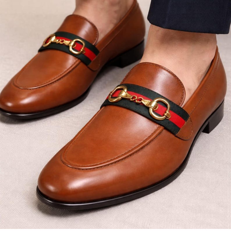 Puede incluir: Mocasines de cuero marr&oacute;n con hebilla dorada y detalle de rayas verdes y rojas. Los zapatos tienen un dise&ntilde;o cl&aacute;sico con punta redondeada y tac&oacute;n bajo.
