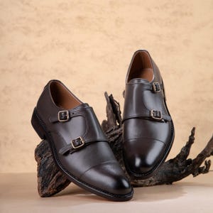 May include: A pair of dark brown leather double monk strap shoes featuring a cap toe design, two adjustable metal buckles on each shoe, and a smooth finish. The shoes are constructed with a low heel and a structured silhouette.