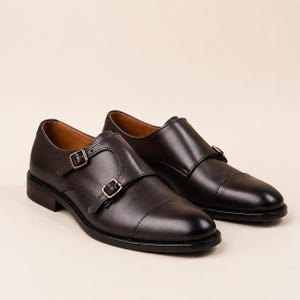 May include: A pair of black leather double monk strap shoes featuring a cap toe design, two silver-toned metal buckles on the side, a low stacked heel, and a smooth finish. The interior lining is a tan color, and the soles are black.