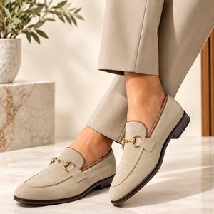 May include: A pair of beige suede loafers featuring a gold-toned metal horsebit detail across the vamp, a brown leather trim along the collar, and a low stacked heel with a dark brown sole, worn with light beige trousers.