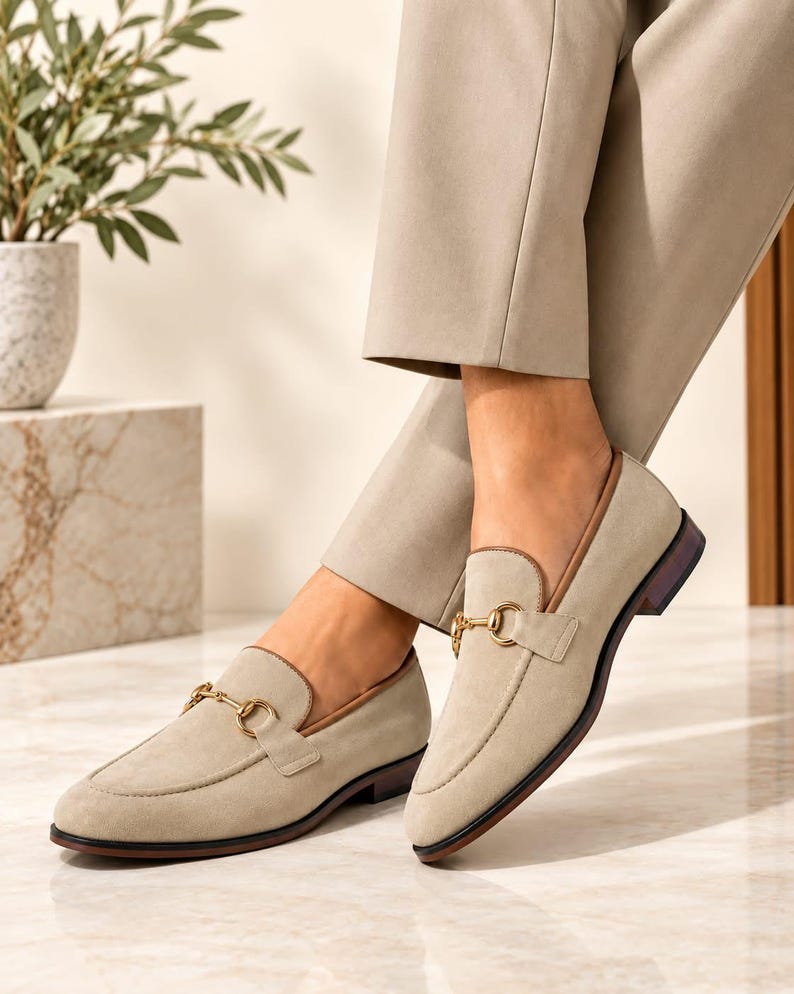 May include: A pair of beige suede loafers featuring a gold-toned metal horsebit detail across the vamp, a brown leather trim along the collar, and a low stacked heel with a dark brown sole, worn with light beige trousers.