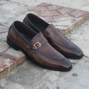 Könnte beinhalten: Ein Paar braune Leder-Loafer mit goldfarbener Schnalle. Die Schuhe haben ein klassisches Design mit runder Spitze und niedrigem Absatz. Das Leder hat eine polierte Oberfläche und die Sohlen sind schwarz.