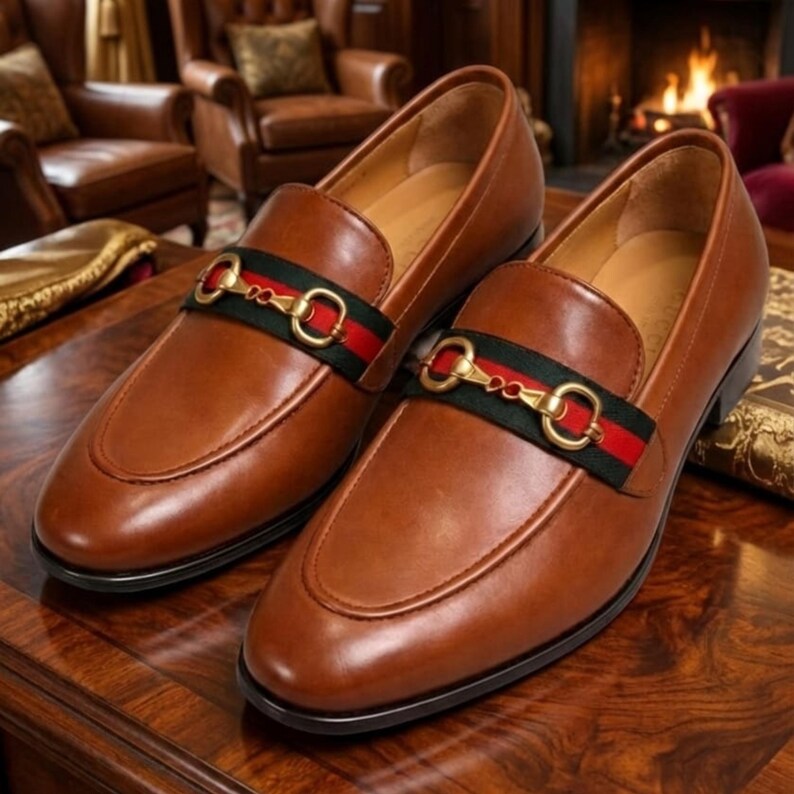 Puede incluir: Un par de mocasines de cuero marr&oacute;n con una correa decorativa con una hebilla dorada y una franja negra y roja. Los zapatos se exhiben sobre una superficie de madera, mostrando su acabado pulido y dise&ntilde;o cl&aacute;sico.