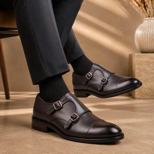 May include: A pair of dark brown leather double monk strap shoes featuring a cap toe design, silver-toned metal buckles, and a low stacked heel. The shoes are worn with black socks and dark trousers, showcasing a polished and smooth leather finish.