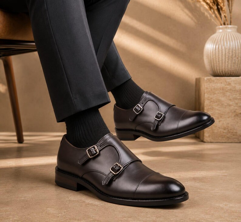 May include: A pair of dark brown leather double monk strap shoes featuring a cap toe design, silver-toned metal buckles, and a low stacked heel. The shoes are worn with black socks and dark trousers, showcasing a polished and smooth leather finish.
