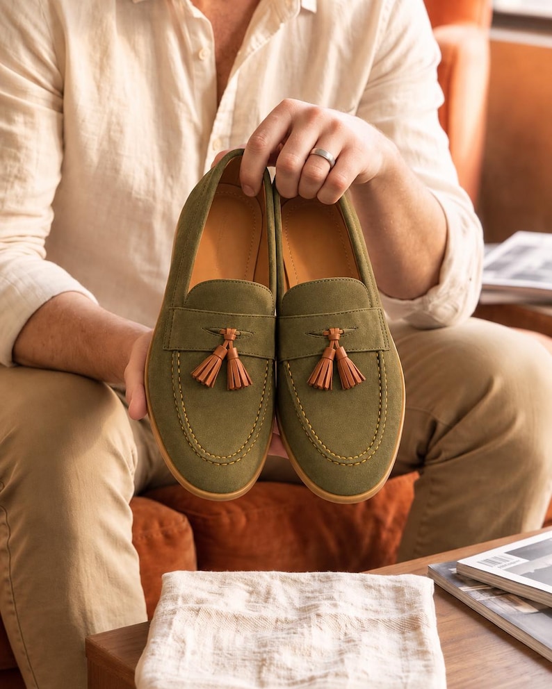 K&ouml;nnte beinhalten: Ein Paar olivgr&uuml;ne Wildleder-Slipper mit Mokassin-Zehenpartie und sichtbaren N&auml;hten. Jeder Schuh ist mit einem dekorativen braunen Lederquasten-Detail &uuml;ber dem Vorderblatt verziert und verf&uuml;gt &uuml;ber ein beiges Innenfutter.