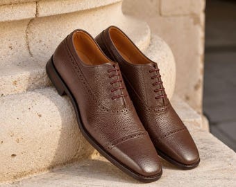 May include: A pair of dark brown leather oxford shoes featuring a textured pebble grain finish, a cap toe design with subtle decorative perforations along the seams, and matching dark brown laces secured through five eyelets on each shoe.