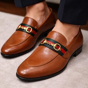 Puede incluir: Mocasines de cuero marr&oacute;n con una correa decorativa con detalles verdes, rojos y dorados. Los zapatos tienen un dise&ntilde;o cl&aacute;sico con punta redondeada y tac&oacute;n bajo. Los mocasines son una elegante opci&oacute;n de calzado.
