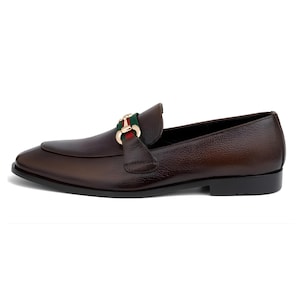 May include: Brown leather loafer with a gold-tone buckle featuring green and red stripes. The shoe has a rounded toe and a low heel. The leather has a textured appearance.