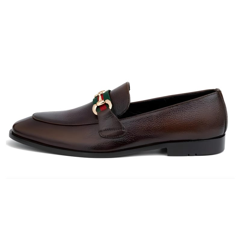 May include: Brown leather loafer with a gold-tone buckle featuring green and red stripes. The shoe has a rounded toe and a low heel. The leather has a textured appearance.