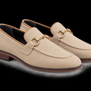 May include: A pair of beige suede loafers featuring a gold-toned metal horsebit detail across the vamp. The shoes have a tan leather trim along the collar, a dark brown stacked heel, and a rounded toe design with visible stitching along the front.