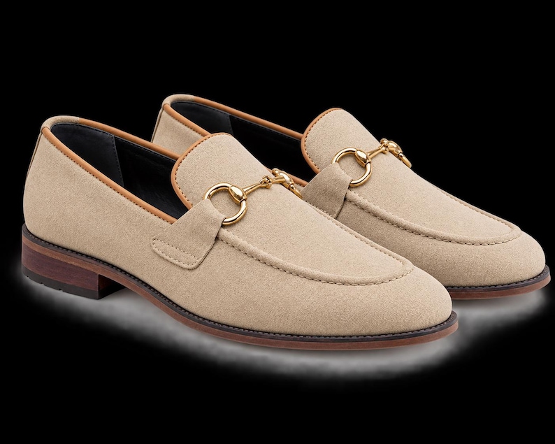 May include: A pair of beige suede loafers featuring a gold-toned metal horsebit detail across the vamp. The shoes have a tan leather trim along the collar, a dark brown stacked heel, and a rounded toe design with visible stitching along the front.