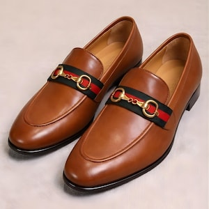Puede incluir: Un par de mocasines de cuero marr&oacute;n con una hebilla de metal dorada y un detalle de rayas verdes y rojas. Los zapatos tienen un dise&ntilde;o cl&aacute;sico sin cordones y un acabado pulido, adecuados para ocasiones formales o semiformales.