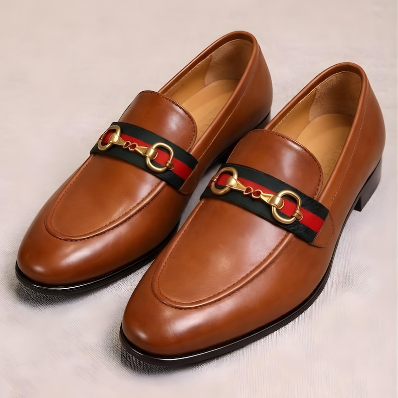 Puede incluir: Un par de mocasines de cuero marr&oacute;n con una hebilla de metal dorada y un detalle de rayas verdes y rojas. Los zapatos tienen un dise&ntilde;o cl&aacute;sico sin cordones y un acabado pulido, adecuados para ocasiones formales o semiformales.
