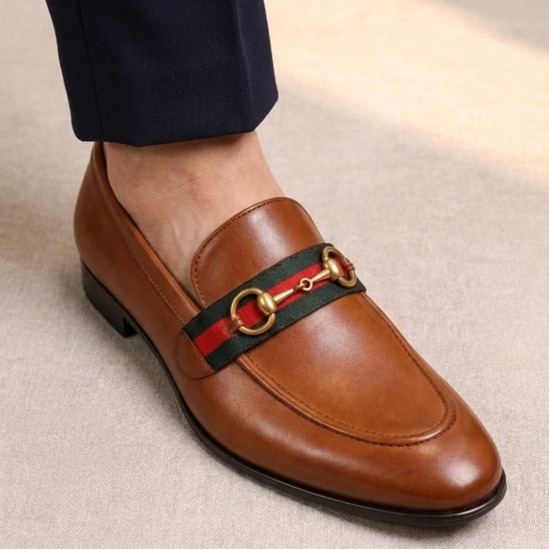 Puede incluir: Mocas&iacute;n de cuero marr&oacute;n con una banda decorativa verde y roja en la parte superior. El zapato presenta una hebilla de metal dorada y una puntera redondeada. El zapato se lleva con un pantal&oacute;n azul marino.