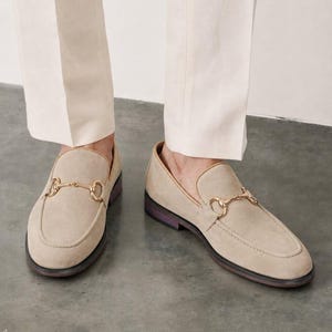 May include: A pair of beige suede loafers featuring a gold-toned metal horsebit detail across the vamp. The shoes have a rounded toe, a low heel, and a dark-colored sole, worn with light-colored trousers that fall just above the ankle.