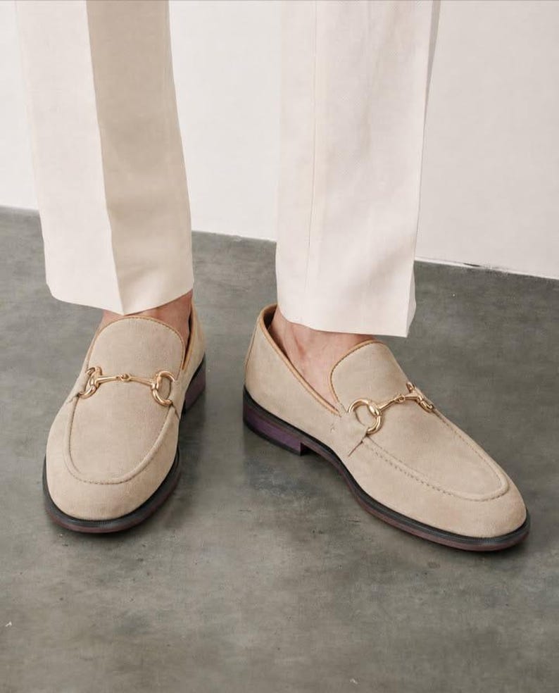 May include: A pair of beige suede loafers featuring a gold-toned metal horsebit detail across the vamp. The shoes have a rounded toe, a low heel, and a dark-colored sole, worn with light-colored trousers that fall just above the ankle.