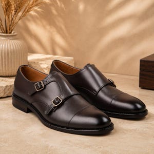 May include: A pair of dark brown leather double monk strap shoes featuring a cap toe design, two adjustable metal buckles on the side, a low heel, and a smooth finish. The interior lining is tan, and the soles are black.