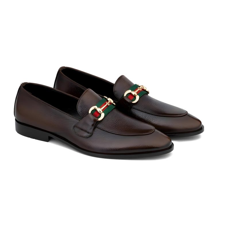 May include: A pair of brown leather loafers with a gold-colored buckle featuring a green and red stripe. The shoes have a classic design with a rounded toe and a low heel. The loafers are displayed on a white background.