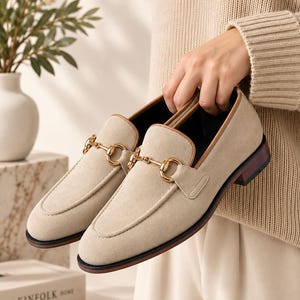 May include: A pair of beige suede loafers featuring a gold-toned metal horsebit detail across the vamp. The shoes have a tan leather trim along the collar, a dark brown stacked heel, and a black outsole, presenting a classic slip-on footwear design.