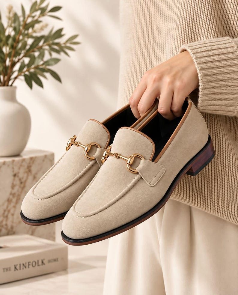 May include: A pair of beige suede loafers featuring a gold-toned metal horsebit detail across the vamp. The shoes have a tan leather trim along the collar, a dark brown stacked heel, and a black outsole, presenting a classic slip-on footwear design.