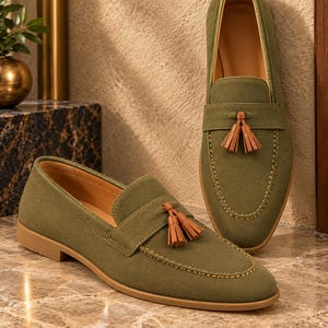K&ouml;nnte beinhalten: Ein Paar olivgr&uuml;ne Slipper mit einem beigefarbenen Lederquasten-Detail auf dem Vorderblatt, einer mokassinartigen Ziernaht an der Schuhspitze und einer flachen beigefarbenen Sohle. Die Schuhe bestehen aus strukturiertem Stoff mit beigem Innenfutter.