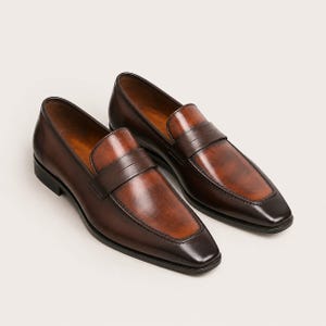 May include: A pair of dark brown leather penny loafers featuring a polished finish with a lighter, burnished tan center panel on the vamp. The shoes have a classic slip-on design with a rounded square toe and a low, stacked heel construction.