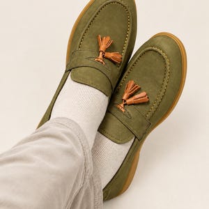 K&ouml;nnte beinhalten: Ein Paar olivgr&uuml;ne Slipper mit glatter Oberfl&auml;che, Mokassin-Naht mit sichtbaren gelben Ziern&auml;hten und einem dekorativen Quasten-Detail aus hellbraunem Leder auf dem Riemen. Die Schuhe haben eine gummierte Sohle und werden mit Socken getragen.