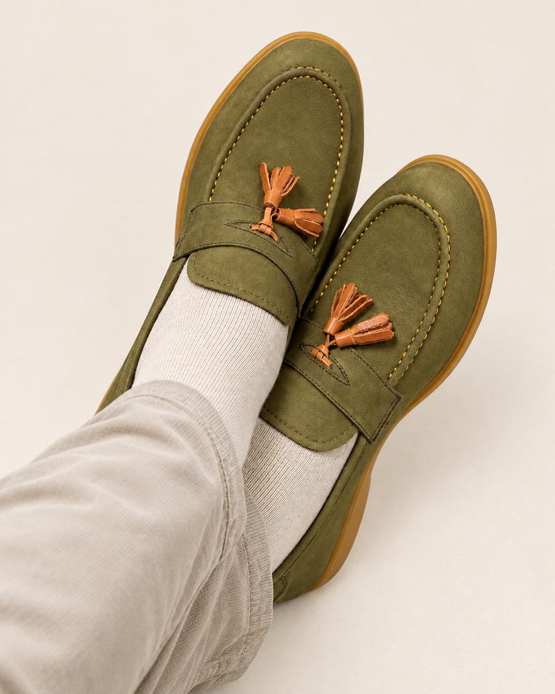 K&ouml;nnte beinhalten: Ein Paar olivgr&uuml;ne Slipper mit glatter Oberfl&auml;che, Mokassin-Naht mit sichtbaren gelben Ziern&auml;hten und einem dekorativen Quasten-Detail aus hellbraunem Leder auf dem Riemen. Die Schuhe haben eine gummierte Sohle und werden mit Socken getragen.