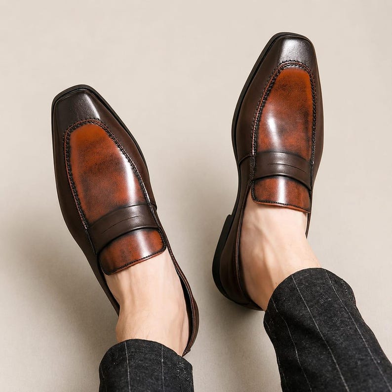 May include: A pair of brown leather loafers featuring a square toe design, a dark brown saddle strap across the vamp, and a burnished finish that transitions from a deep dark brown at the edges to a lighter reddish-brown tone in the center.