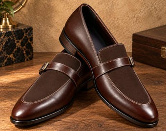 May include: A pair of dark brown leather and suede loafers featuring a single monk strap with a metallic buckle. The shoes have a smooth leather outer frame, a textured suede vamp, and a low black heel with visible white contrast stitching.
