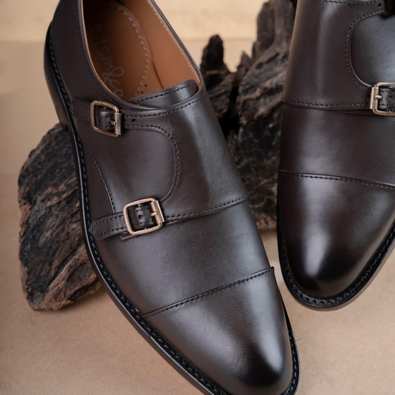 May include: A pair of dark brown leather double monk strap shoes featuring a cap toe design, two adjustable metal buckles on the side, and a smooth finish. The interior lining is tan with a visible brand logo printed on the inner sole.