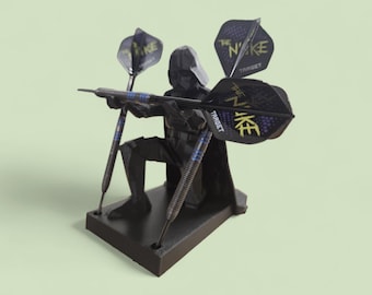 Könnte beinhalten: Ein schwarzer Dart-Halter in Form einer knienden Figur, die Darts hält. Die Darts haben schwarze und goldene Flights mit dem Text "The Nuke Target". Der Halter steht auf einem rechteckigen Sockel mit hellgrünem Hintergrund.