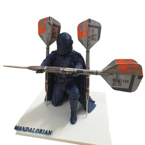 May include: A dark blue Mandalorian figurine kneeling and holding a dart. The figure is positioned in front of three darts with orange and grey wings. The base of the figurine reads "MANDALORIAN".
