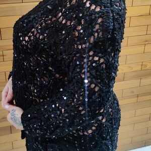 May include: A black, open-weave top with long sleeves, heavily embellished with sequins. The top has a loose fit. It is worn over a light-coloured garment. The background is a wooden wall.